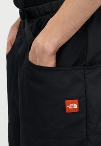 Τα χέρια ενός ατόμου στις τσέπες μαύρου παντελονιού North Face με πορτοκαλί λογότυπο κοντά στην τσέπη στο δεξί πόδι.