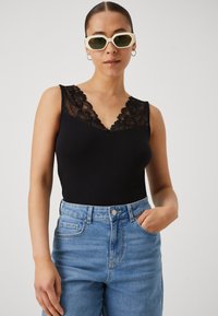 Bodysuit sans manches noir avec des détails en dentelle au niveau du col, associé à un jean taille haute bleu clair. Modèle portant des lunettes de soleil teintées en vert.