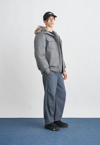 Graue Daunenjacke mit einer mit Pelz besetzten Kapuze, zwei Brusttaschen und Rippbündchen, kombiniert mit lockeren blauen Hosen und schwarzen Sneakers.