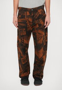 Braune Hose mit einem Muster aus verschiedenen gedruckten Grafiken, aus einem weichen Stoff, mit einer lockeren Passform und zwei Fronttaschen.