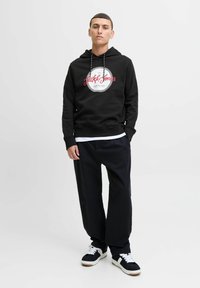 Sort hætte-trøje med hvidt og rødt logo, fremstillet af tekstureret stof. Kombineret med løstsiddende marineblå bukser og sorte sneakers.