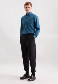 Blågrøn knap-op skjorte med lange ærmer, brystlomme og hvide knapper, kombineret med sorte plisserede bukser og sorte pæne sko.