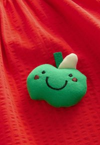 Roter strukturierter Stoff mit einem applizierten, apfelförmigen Patch in Grün. Der Patch zeigt ein lächelndes Gesicht, mit gestickten Details und einem kleinen weißen Blattakzent.