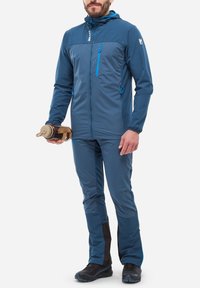 Veste légère bleue avec capuche, poches zippées et fermetures éclair bleues contrastantes, associée à un pantalon bleu assorti et des chaussures noires.