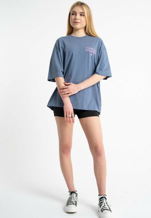 Młoda kobieta stoi z założonymi ramionami, ubrana w luźny niebieski T-shirt z napisem „Thank You Being Kind”, czarne szorty i trampki.