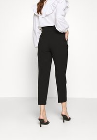 Svarta skräddarsydda byxor med smal passform, hög midja och sidofickor, ihopparat med en vit blus med volanger och svarta klacksandaler.