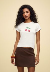 Vitt kortärmat t-shirt med ett körsbärsgrafik i röd och grön. Kombineras med en brun kjol. Modellen bär färgglada pärlarmband.