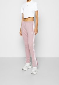 Rosa polyester-joggingbyxor med vita sidostrimlor och dragkedjor vid vristen, i kombination med en vit kort topp med en liten logotyp.