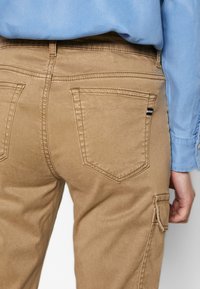 Jasnobrązowe spodnie cargo o gładkiej teksturze, z zakrzywionymi tylnymi kieszeniami i kieszenią boczną, zestawione z niebieską koszulą.