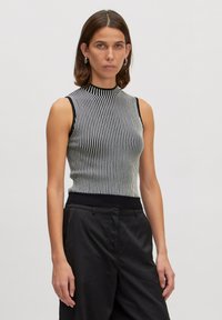 Schwarzes und weißes gestreiftes ärmelloses Oberteil mit hohem Kragen und geripptem Textil; hat ein schwarzes Band an der Taille. Getragen mit schwarzen Hosen.