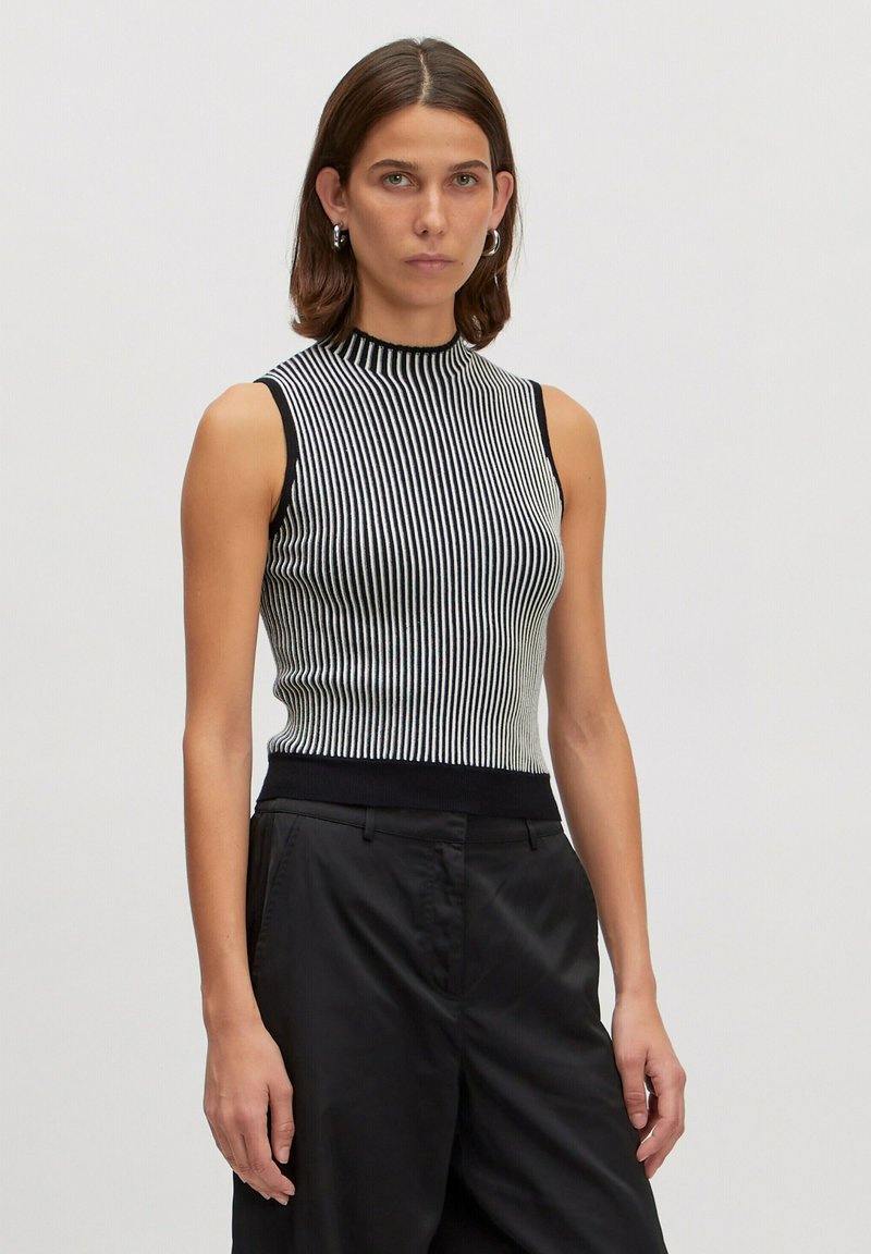 Zwart-wit gestreepte mouwloze top met een hoge halslijn en ribbeltextuur; heeft een zwarte band in de taille. Draagt met zwarte broek.