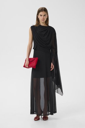 Γυναίκα που φοράει μαύρο φόρεμα χωρίς μανίκια με διαφανές επάνω στρώμα και λεπτομέρειες draped, κρατώντας κόκκινη τσάντα χειρός και φορώντας κόκκινες σανδάλες.