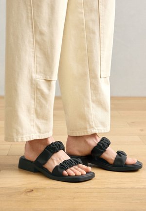 Des sandales noires avec des sangles à volants rembourrées, une semelle plate et un design à bout carré, associées à des pantalons larges beige clair sur un sol en bois.