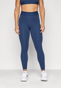 Leggings deportivos de color azul marino con una cinturilla gruesa, textura suave y detalle de logotipo. Combinados con zapatillas blancas.