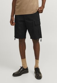 Sorte cargo shorts med sidelommer, snoretræk i kanten og en afslappet pasform, parret med beige ribbede strømper og sorte lædersko.