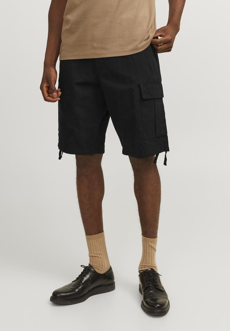 Sorte cargo shorts med sidelommer, snoretræk i kanten og en afslappet pasform, parret med beige ribbede strømper og sorte lædersko.