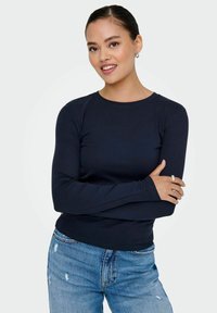 Camiseta de manga larga en diseño ajustado, hecha de un tejido suave. Combinada con jeans desgastados de lavados claros. El modelo se encuentra de pie con los brazos cruzados.