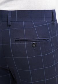 Navy plaid trousers featuring a smooth fabric texture, a rear pocket with a button closure, and a tailored fit design.