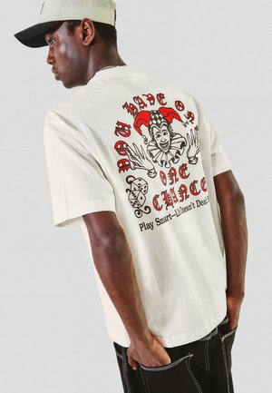 Młody mężczyzna w białej czapce i kremowej koszulce z grafiką błazna, kostkami do gry i tekstem „Masz tylko jedną szansę” oraz „Grać mądrze – życie nie jest sprawiedliwe.”