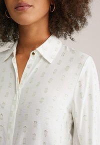Camicia bianca con bottoni, caratterizzata da un tessuto testurizzato con motivi metallici verdi. Ha un colletto classico e sottili accenti brillanti.