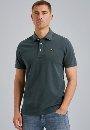 Donkergroene polo gemaakt van katoen, met een drieknoopsluiting en een klein gouden embleem op de linkerborst. Korte mouwen.