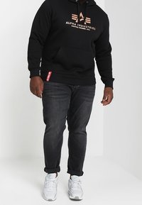 Felpa nera con cappuccio e tasca frontale, con logo "Alpha Industries" in bronzo. Abbinata a jeans scuri e sneakers bianche. Vestibilità casual.