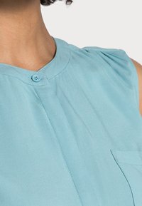 Blouse sans manches bleu clair avec un col arrondi, fermeture par boutons et une poche avant ; dotée d'un tissu doux avec une texture subtile et un détail plissé.