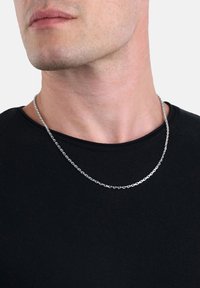 Närbild av en mans nedre ansikte och nacke som bär ett silverhalsband över en svart T-shirt med rund halsringning.