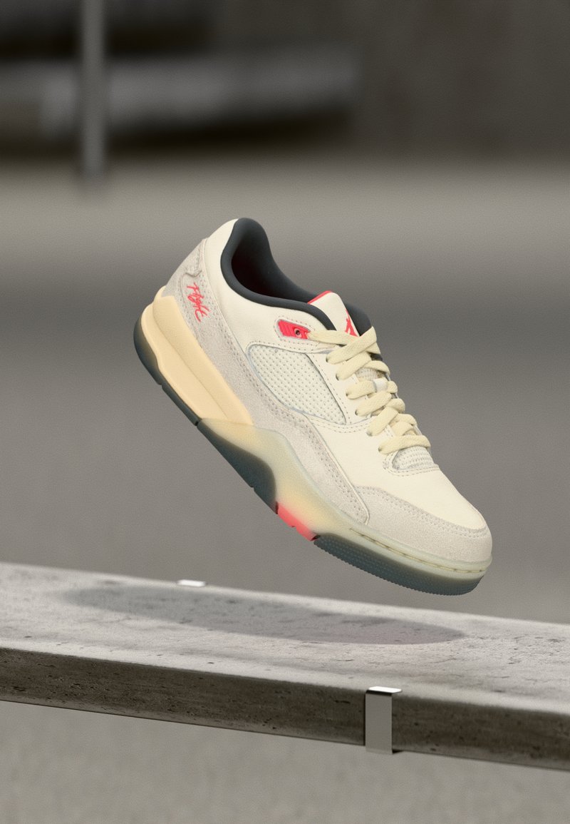 Κρέμα και μπεζ sneaker με κόκκινες λεπτομέρειες που αιωρείται πάνω από μια τσιμεντένια ακμή, δείχνοντας την υφασμένη σόλα και τις ραμμένες λεπτομέρειες.