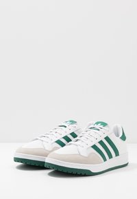 Sneakers with a white fabric upper, green suede accents, and three green stripes. Features a textured toe and a padded collar.