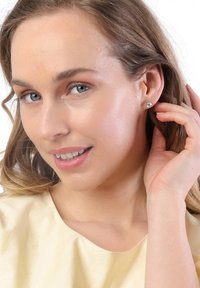 Modèle portant de petites boucles d'oreilles claires rondes, avec de longs cheveux ondulés. Haut jaune clair, texture douce, maquillage naturel, arrière-plan neutre.