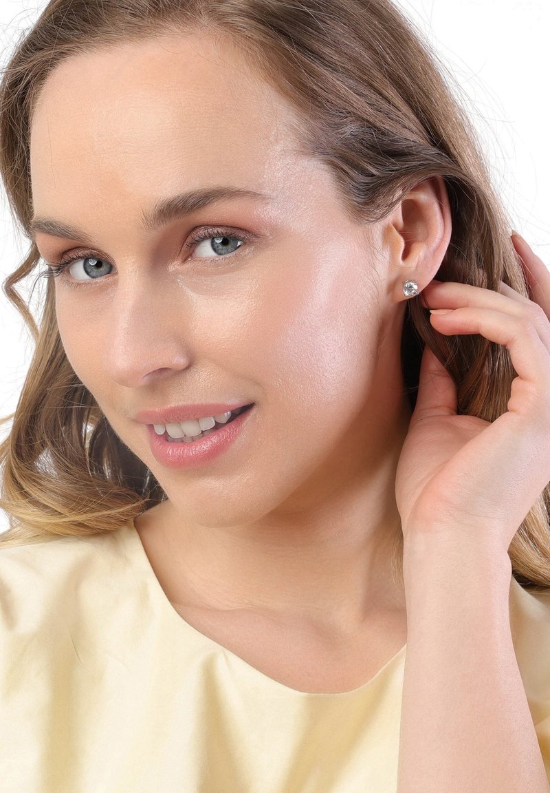 Modèle portant de petites boucles d'oreilles claires rondes, avec de longs cheveux ondulés. Haut jaune clair, texture douce, maquillage naturel, arrière-plan neutre.