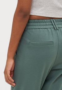 Grønne, elastiske bukser laget av stoff med en elastisk midjebånd. En liten, horisontal baklomme er synlig nær midjen.