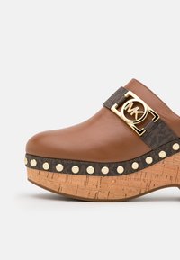 Mule compensée en cuir marron avec plateforme en liège, clous dorés, bordure à motif sombre, et boucle logo Michael Kors dorée sur la bride latérale.