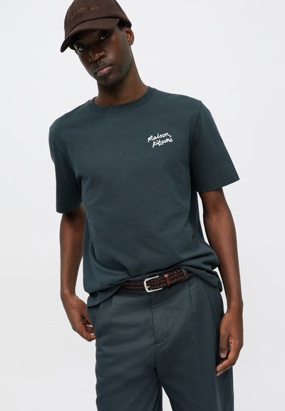 Dunkelgrünes Baumwoll-T-Shirt mit Rundhalsausschnitt und kurzen Ärmeln, mit weißem gesticktem Text auf der Brust. Kombiniert mit dunklen Hosen und einem braunen Gürtel.
