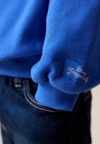 Sweatshirt bleu avec des poignets côtelés, portant une étiquette rose "Disney Princess" cousue sur la manche gauche. Porté avec un jean foncé à coutures visibles.