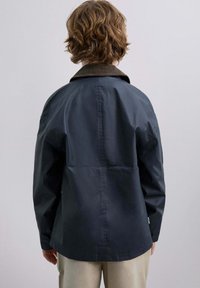 Persona con cabello ondulado que lleva una chaqueta azul oscuro con un cuello marrón, vista desde atrás contra un fondo liso de color gris claro.