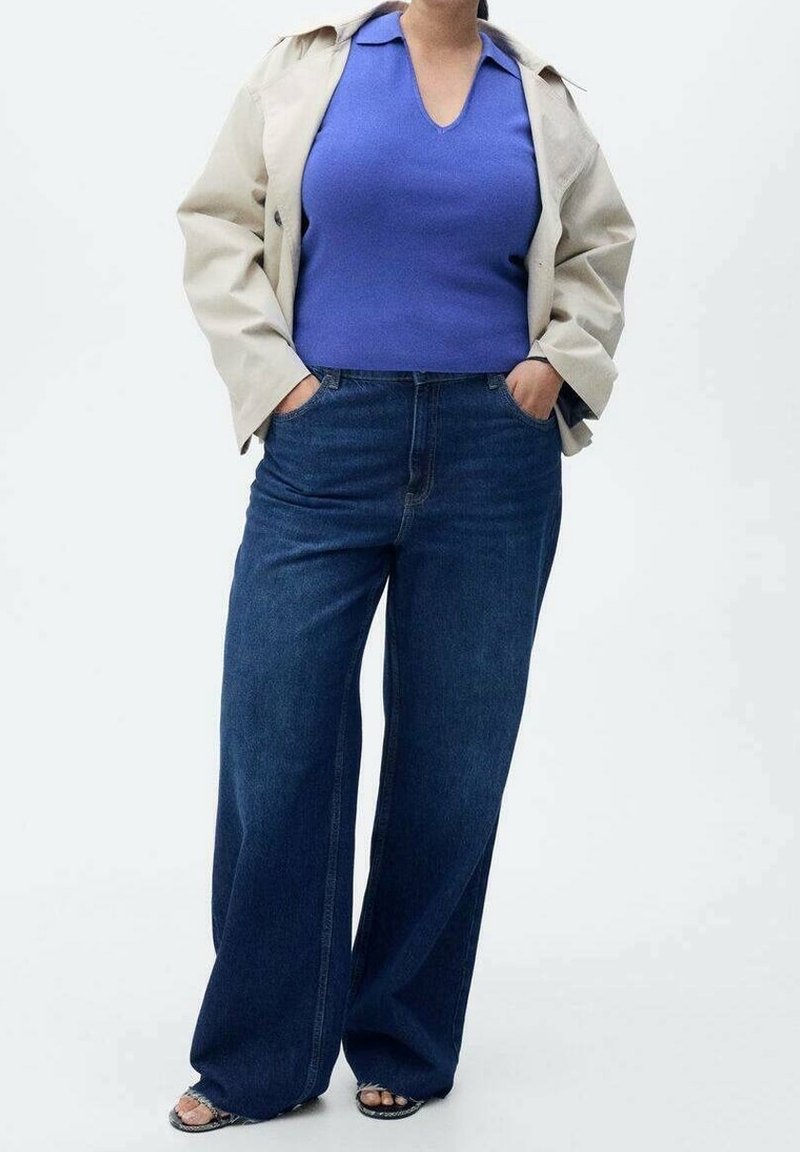 Personne portant une veste beige, un haut sans manches bleu, un jean large bleu foncé et des sandales à bouts ouverts, debout les mains dans les poches.