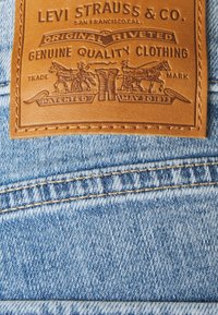 Ruskea nahkainen merkki, jossa on kohokuvioitu teksti ja kuvitus, kiinnitetty vaaleansiniseen farkkukankaaseen. Merkissä on haalistunut, teksturoitu pinta ja kontrastiväriset saumat.