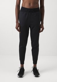 Schwarze sportliche Jogginghosen mit geripptem Bund, Kordelzug, zwei Seitentaschen und schmal zulaufenden Beinabschlüssen. Getragen mit schwarzen Laufschuhen.