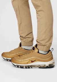 Személy arany Nike Air Max 97 sneakerben, bézs nadrágban és fehér zokniban, fehér háttér előtt.