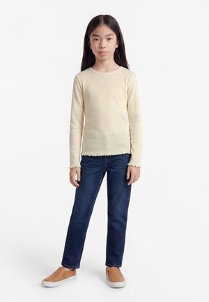 Young girl with long straight hair wearing a cream patterned long-sleeve top, dark blue jeans, and tan slip-on shoes, standing facing forward.