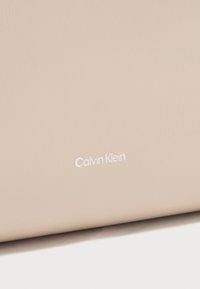 Calvin Klein bežasti teksturirani materijal s glatkom finišom; sadrži bijeli urezani logo. Jednostavan dizajn bez dodatnih uzoraka ili naglasaka.
