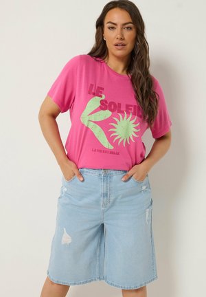 Kvinde med langt mørkt hår iført en pink T-shirt med grafik og lyseblå slidte cowboyshorts til knæet med hænderne i lommerne.