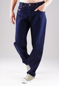 Marineblaue Jeans mit lockerer Passform, ausgestattet mit kupferfarbenen Beschlägen und einem Fünf-Taschen-Design. Kombiniert mit grünen und weißen Sneakern.