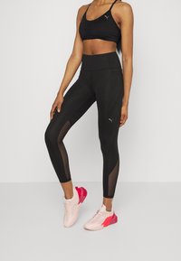 Un sujetador deportivo negro y leggings de cintura alta presentan un patrón texturizado y paneles de malla. El calzado es de color rosa claro con una suela rosa.