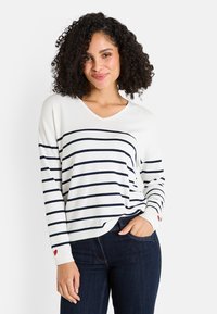 Mujer con cabello rizado que lleva un suéter blanco de manga larga con rayas horizontales en azul marino y pequeños acentos en forma de corazón rojo, combinado con jeans de color azul oscuro.