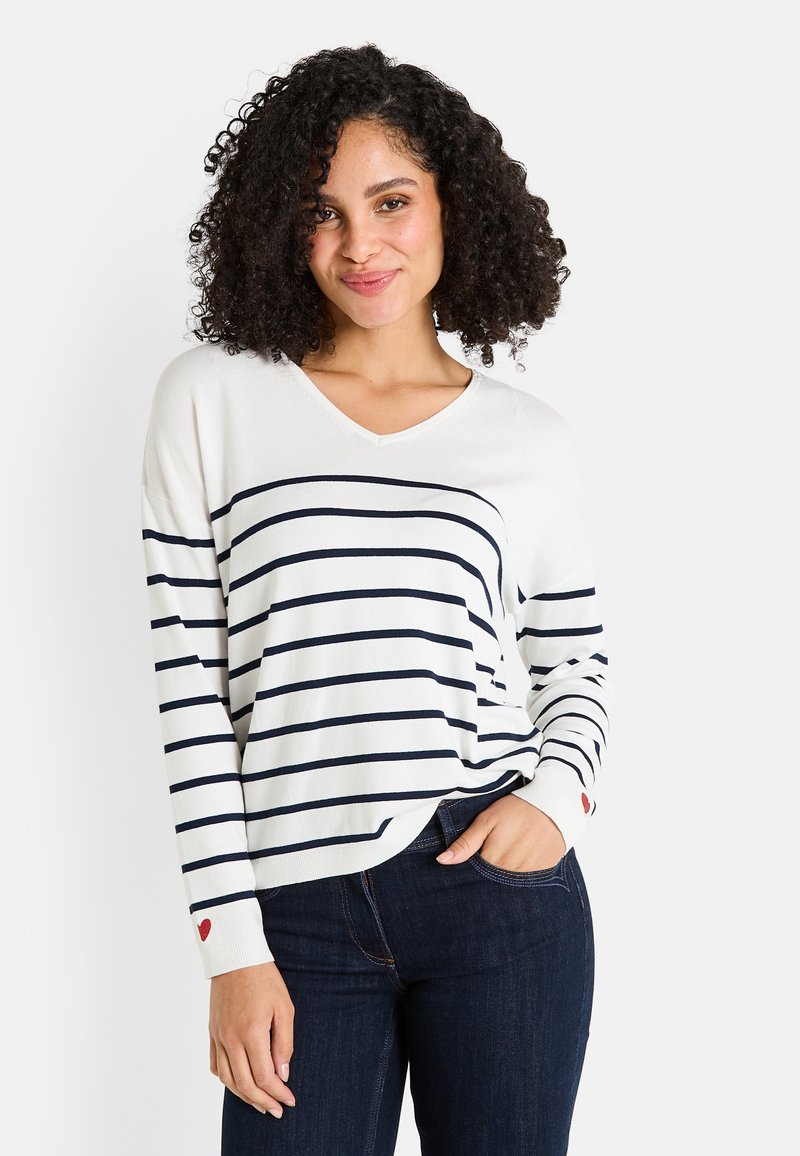 Mujer con cabello rizado que lleva un suéter blanco de manga larga con rayas horizontales en azul marino y pequeños acentos en forma de corazón rojo, combinado con jeans de color azul oscuro.