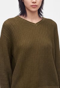 Femme portant un pull en maille vert olive texturé avec col en V, montrée des épaules au milieu du torse sur fond uni.