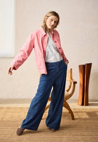 Femme se penchant sur le côté, portant une veste rose, une chemise blanche à motifs, un pantalon large bleu et des chaussures marron dans une pièce minimaliste avec une chaise en bois et une décoration.